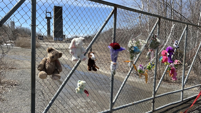 Des peluches et des fleurs accrochés sur une clôture qui ferme un site industriel désaffecté.