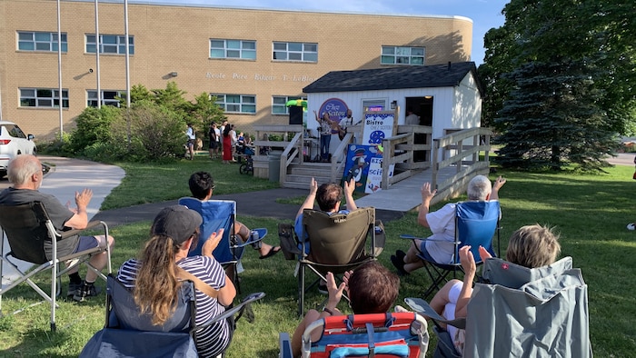 Des clients assis sur des chaises de camping applaudissent un spectacle de musique devant le bistro.