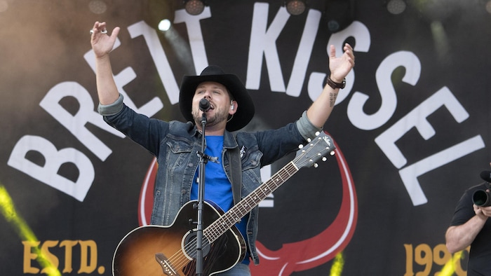 Un chanteur devant son micro est coiffé d'un chapeau de cowboy noir, a une guitare au cou et lève les bras au ciel.