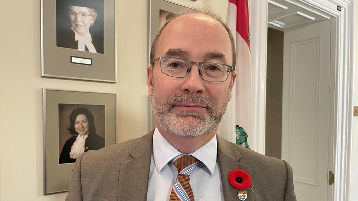 Matt MacFarlane regarde directement la caméra. Il est debout contre un mur où sont accrochés des portraits de femmes en toge.