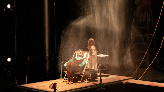 En scène de la pièce de théâtre «Créatures» où une petite fille est debout aux côtés d'une femme assise dans une chaise sous une good bruine d'eau.