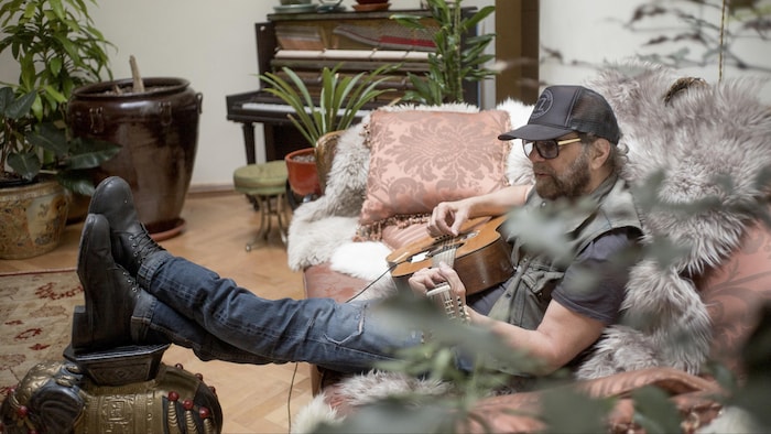 Un homme qui joue de la guitare, assis sur un sofa.
