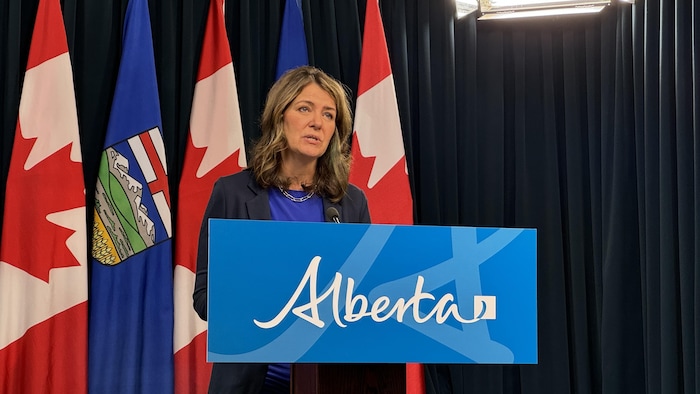 Danielle Smith debout devant un lutrin avec des drapeaux de l'Alberta et du Canada derrière elle.