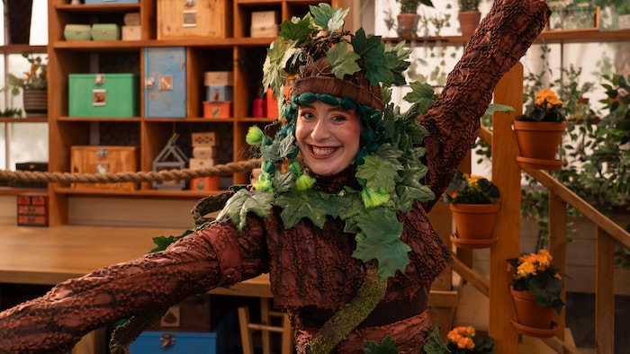 Une femme souriante en costume d'arbre qui pose dans un décor rempli de plantes.