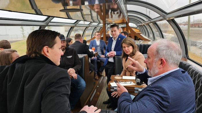 Devin Dreeshen, David Moretta et Danielle Smith discutent dans un wagon dont le toit est entièrement vitré. D'autres personnes sont présentes dans le wagon. 