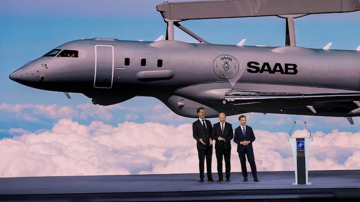 Trois hommes devant un jet militaire sur une scène lors d'un événement.