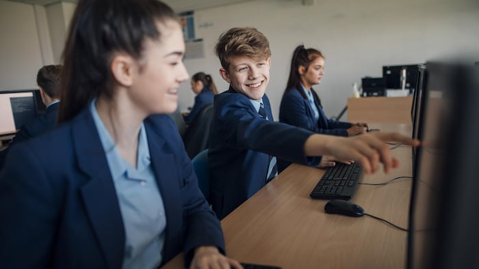Des élèves qui sont devant des ordinateurs dans une salle de classe. Un d'eux pointe vers un écran.