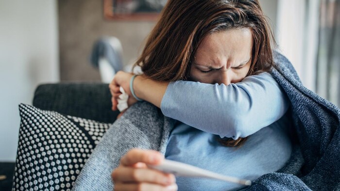 Une femme tousse dans son coude gauche. Elle tient un thermomètre dans la main droite.