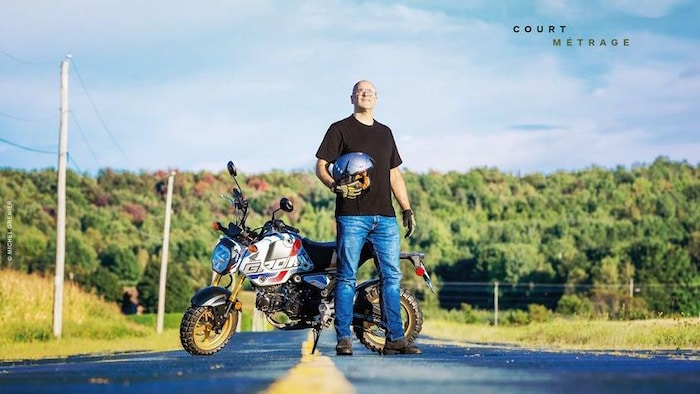 Daniel Grenier est devant une moto sur la route. Guimauve est écrit dans le ciel. 