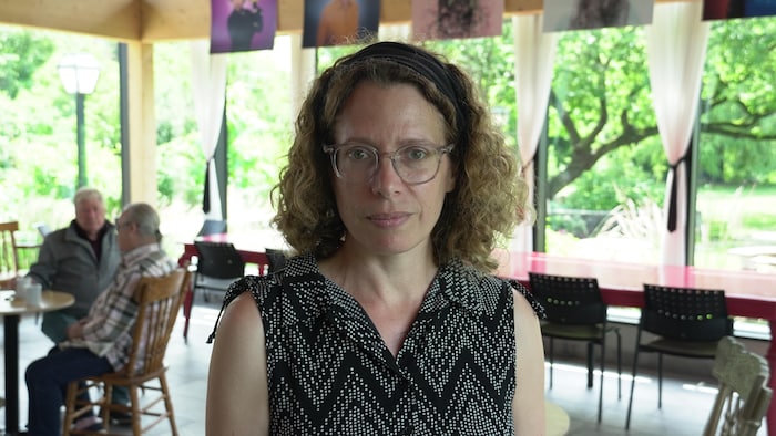 Une femme aux cheveux frisés fixe l'objectif. Elle est debout dans la verrière du Théâtre de l'île. En arrière-plan, connected aperçoit, deux hommes en treatment autour d'un café.