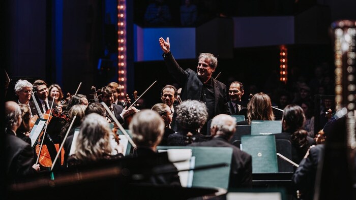 Le chef d'orchestre John Storgårds lève un bras en l'air, souriant, entouré de musiciens d'un orchestre.