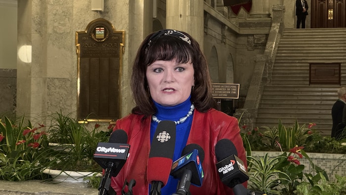 Kris Sims parle devant plusieurs micros devant les grands escaliers de l'Assemblée législative de l'Alberta.