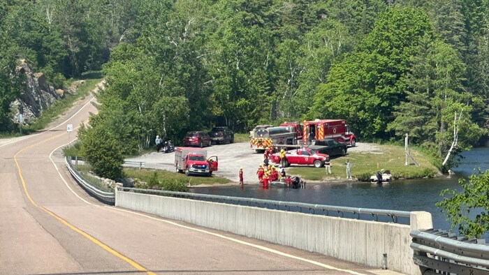 Des véhicules et du personnel du service de sécurité incendie en bordure d'un lac et d'une route dans une forêt.