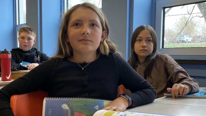 Une élève est assise à son pupitre. À sa gauche et derrière elle, il y a une autre fille et un garçon.