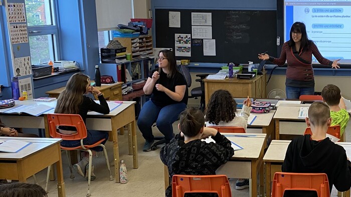 À droite, une enseignante parle devant une classe. À gauche, une interprète traduit en langue des signes à une élève sourde.
