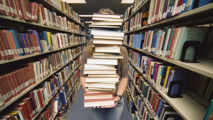 Une femme qui tient une grande pile de livres devant elle. Elle est dans une rangée d'une bibliothèque.