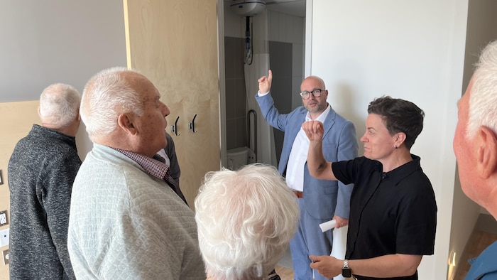 Des personnes âgées regardent une personne parler, qui pointe quelque chose du doigt.