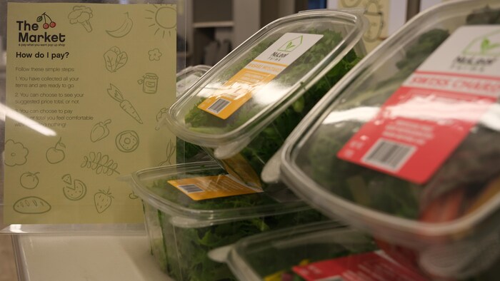 Des boîtes de salades sont alignées sur une table. Au loin, une affiche explique le fonctionnement du marché. 
