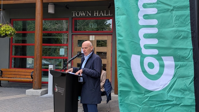 Marco Viviani parle à un lutrin devant la mairie de Banff. Une bannière de l'entreprise flotte sur le côté.