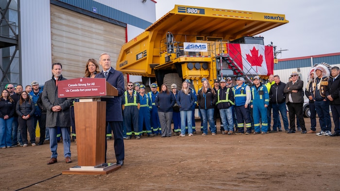 Mark Carney parle à un podium, avec Danielle Smith derrière lui, ainsi que des travailleurs de l'industrie pétrolière, lors d'une annonce à Fort McMurray le 1er octobre 2026.