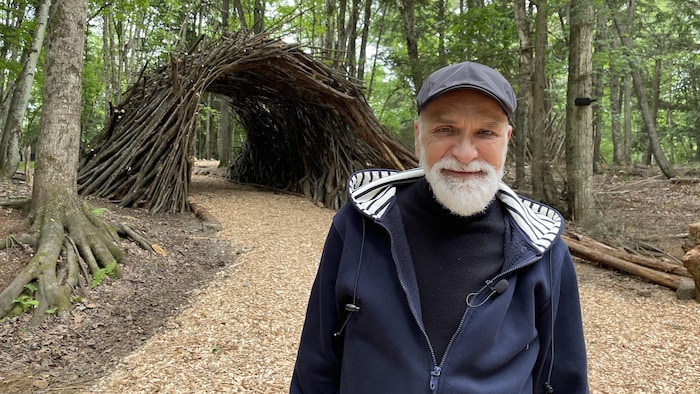 Un homme souriant devant une structure faite de branches de bois dans une forêt.