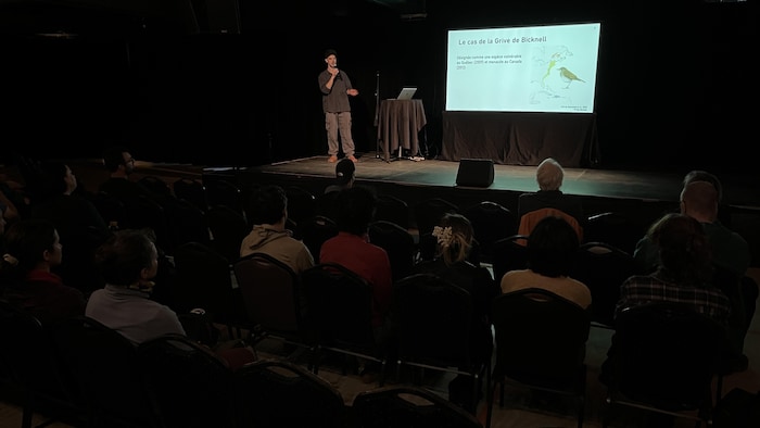 Alexandre Ethier est sur un scène. Il parle dans un microphone en faisant face à plusieurs personnes assises sur des chaises. Un support visuel est projeté.