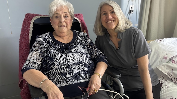 Janita Richard est assise dans sa chaise, aux côtés de son lit. Isabelle Moisan est à côté d'elle. Les deux regardent la caméra en souriant. 