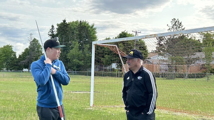 Jeune lanceur de javelot discute avec son entraîneur devant un filet de soccer.