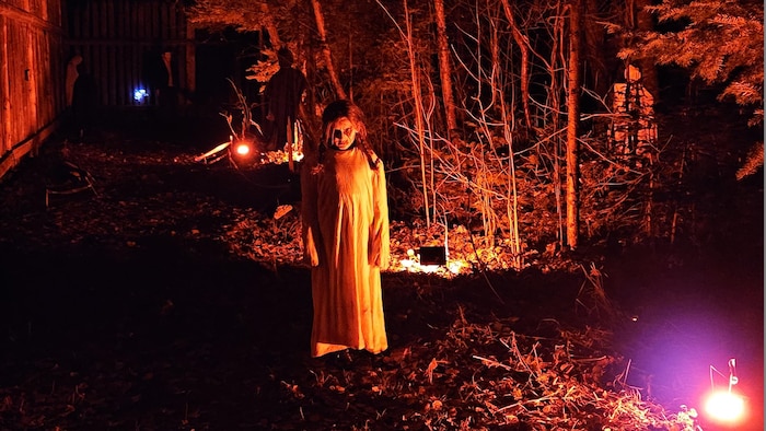 Une comédienne en zombie vêtue d'une longue robe blanche se tient dans les bois éclairés par des projecteurs rouges.