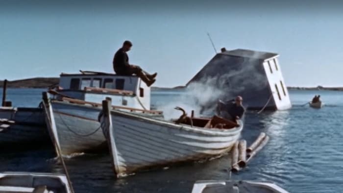 Une maison flotte sur l'eau et est tirée par trois petits bateaux à moteur.