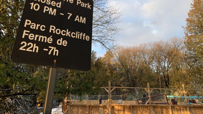 En avant-plan, un panneau affichant les heures d'ouvertures d'un parc. En arrière-plan, une patinoire dans une forêt.