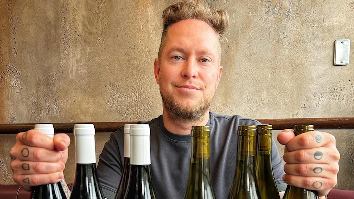 Un homme blond derrière plusieurs bouteilles de vin.