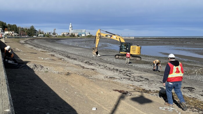Une pelle mécanique sur une plage, où se trouvent plusieurs travailleurs de la construction.