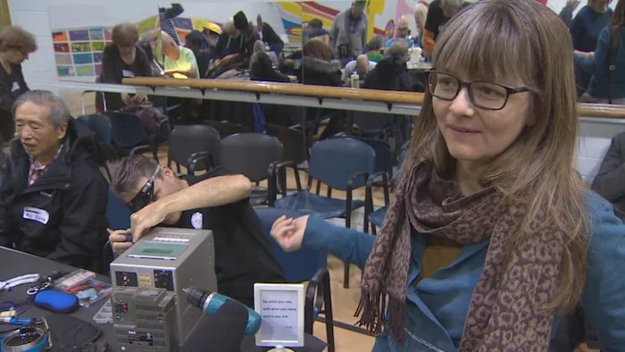 Une femme se trouve dans un café de réparation. Au deuxième plan, un homme répare un appareil électronique.
