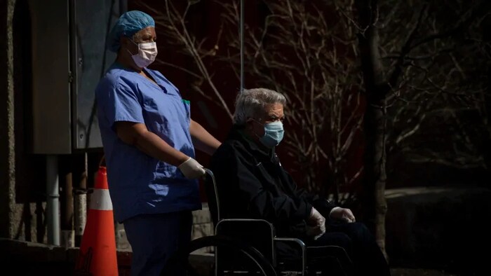 Une employée d'une résidence de soins de longue durée pousse un patient assis sur une chaise roulante. Les deux personnes sont masquées.