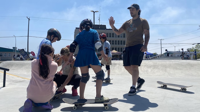 Une maman et sa fille aide son jeune fils, Oli, à tenir debout sur une planche à roulettes. Juste à côté, un moniteur s'approche pour donner un « high five » d'encouragement.