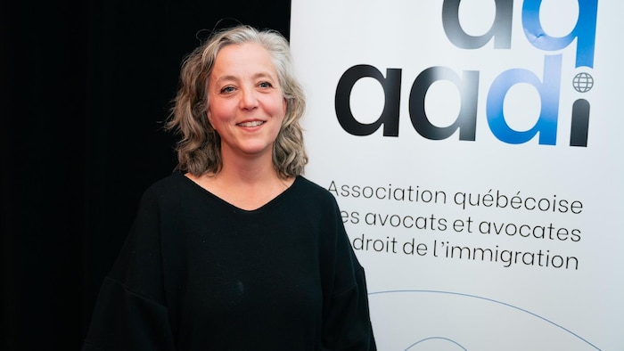 Une femme souriante aux côtés d'une bannière de l’Association québécoise des avocats et avocates en droit de l’immigration.