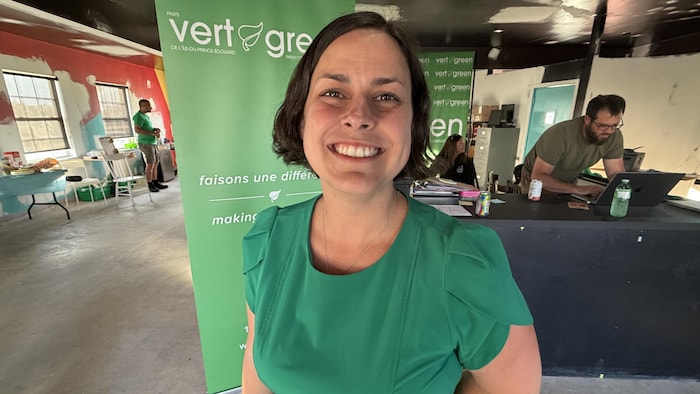 Une femme aux cheveux courts foncés, toute de vert vêtue, se tourne vers la caméra et fait un large sourire.