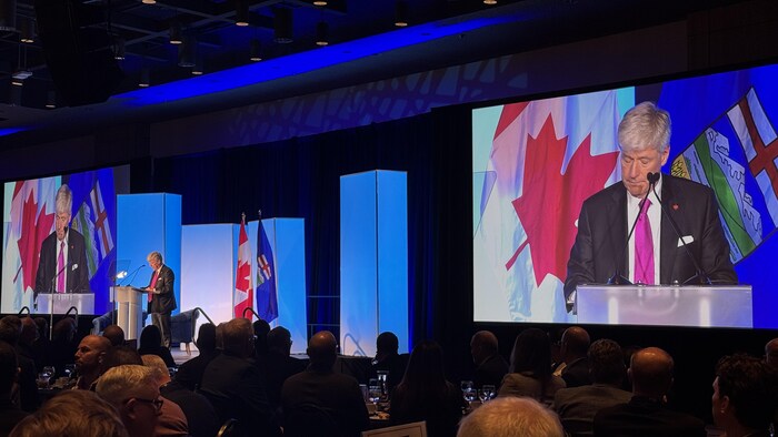 Tim Hodgson parle devant un vaste public. Il est entouré de deux écrans géants et des drapeaux du Canada et de l'Alberta.