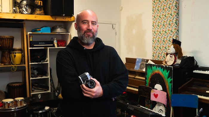 Un homme souriant qui pose devant des instruments de musique. Il tient un appareil photo.
