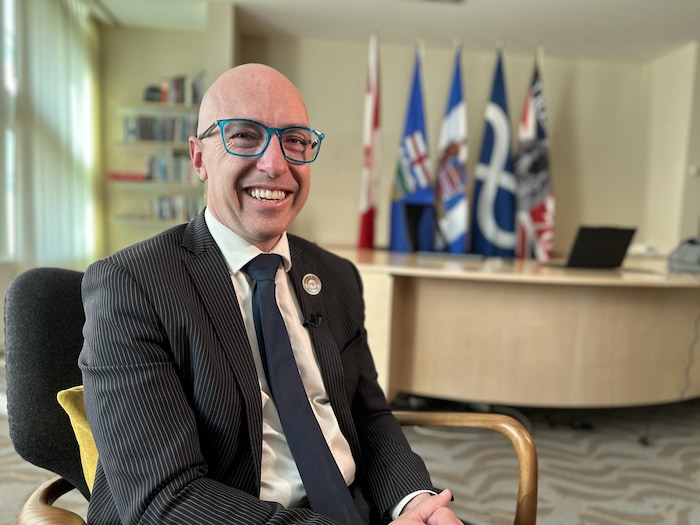 Un homme chauve et souriant en complet sombre avec des lunettes bleues est assis dans un bureau avec des drapeaux flous en arrière-plan, le 28 octobre 2025.