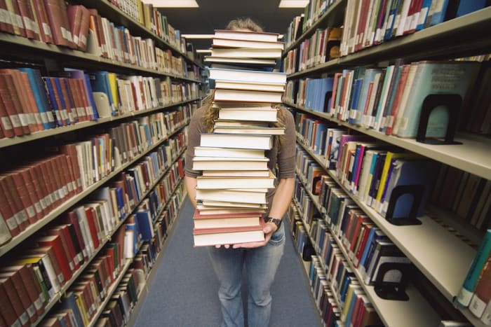 Une femme qui tient une grande pile de livres devant elle. Elle est dans une rangée d'une bibliothèque.