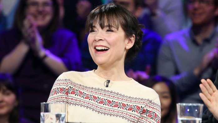 Elle porte un chandail à petit imprimé de fleurs. Elle sourit sur un plateau de télévision. On voit du public derrière elle en studio.