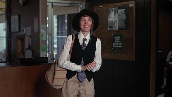 Une femme, Diane Keaton, porte une cravate et un chapeau en souriant.