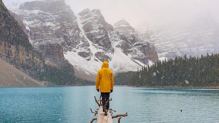 Une personne avec un manteau jaune est debout sur un tronc d'arbre au bord de l'eau et il neige.