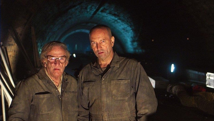 Deux hommes (Jean Lapointe et Michel Côté) en combinaison de travail, dans un tunnel.