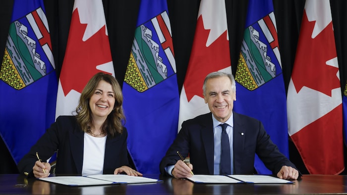 Une femme et un homme très souriants tenant chacun un crayon, devant une rangée de drapeaux du Canada et de l'Alberta.
