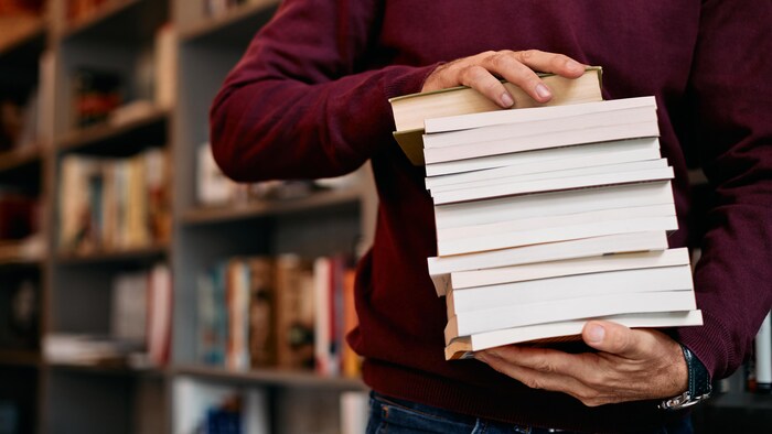 Dans les rangées d'une bibliothèque, un homme tient une pile de livres entre ses mains.