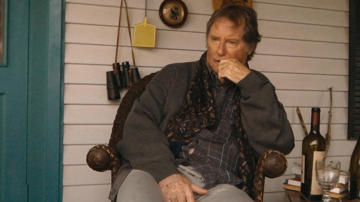Pop (Marc Messier) assis sur sa galerie. Il y a deux bouteilles de vin et un verre sur une petite table à côté de lui.