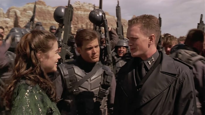 Une jeune femme parle à un jeune soldat, entouré d'autres hommes en tenues de combat.